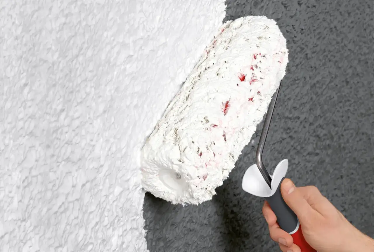 Dłoń trzyma wałek do malowania, który pokrywa teksturalną ścianę białą farbą. Dowiedz się, jak wybrać wałek do malowania.