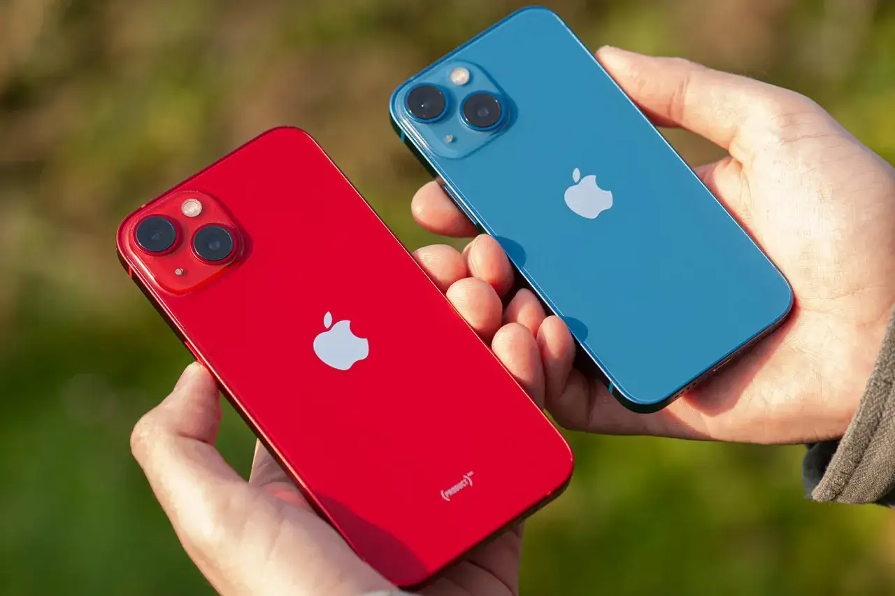 Dwie ręce trzymają dwa telefony: czerwony i niebieski iPhone 13 mini. Ich aparat jest gotowy do robienia zdjęć.