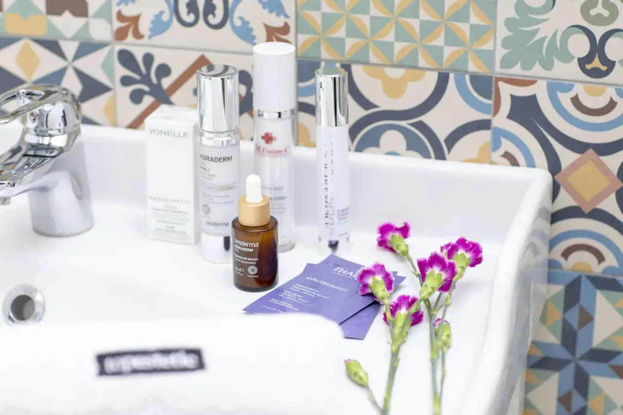 Kosmetyki z kwasem hialuronowym, w tym serum i kremy, leżą w białej umywalce obok kolorowych płytek i kwiat&oacute;w.