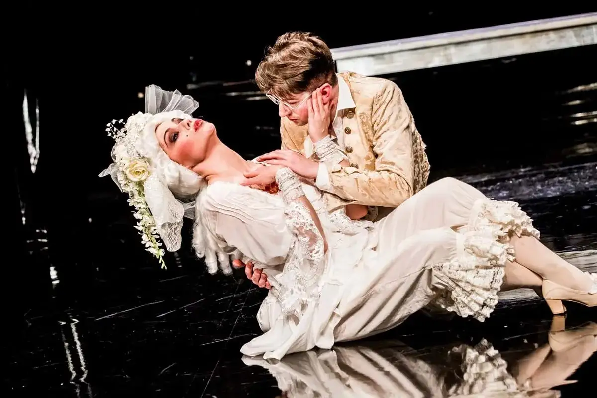 Sylwia Janowicz-Dobrowolska w białej sukni i peruce, obejmowana przez mężczyznę w złotej kurtce, na czarnej scenie.