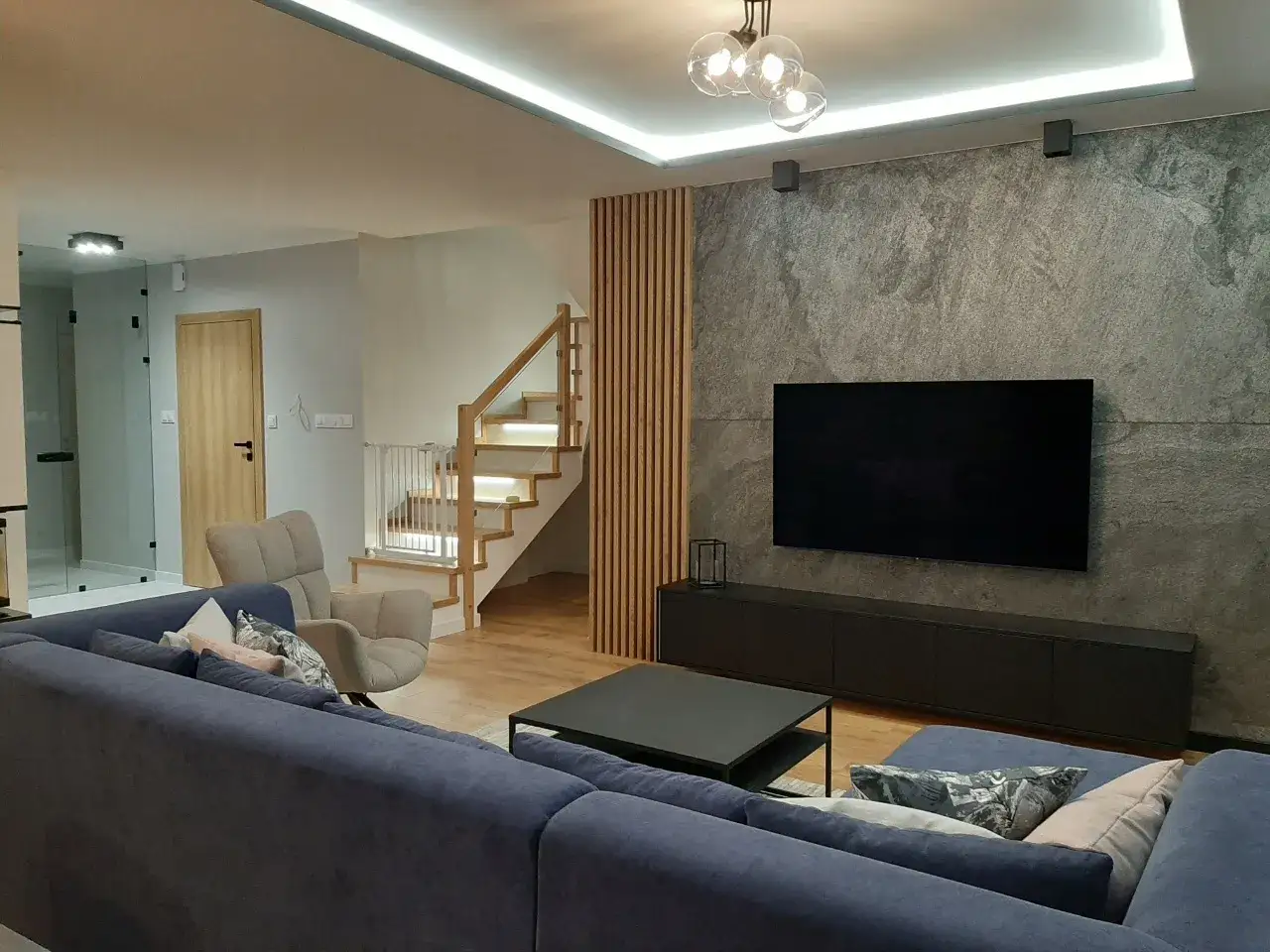 Nowoczesna aranżacja ściany przy schodach z kamiennym wykończeniem i telewizorem. Drewniane panele dodają ciepła.