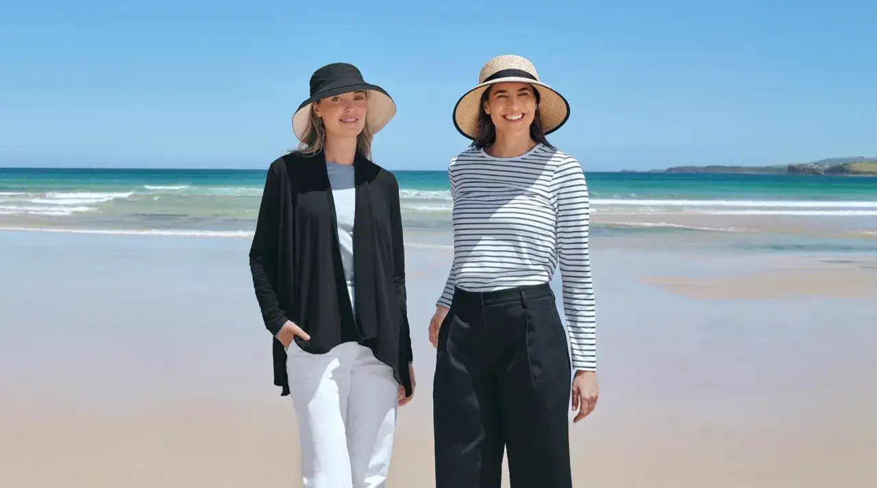 Dwie kobiety na plaży, obie w stylowych kapeluszach zapewniających ochronę głowy przed słońcem.