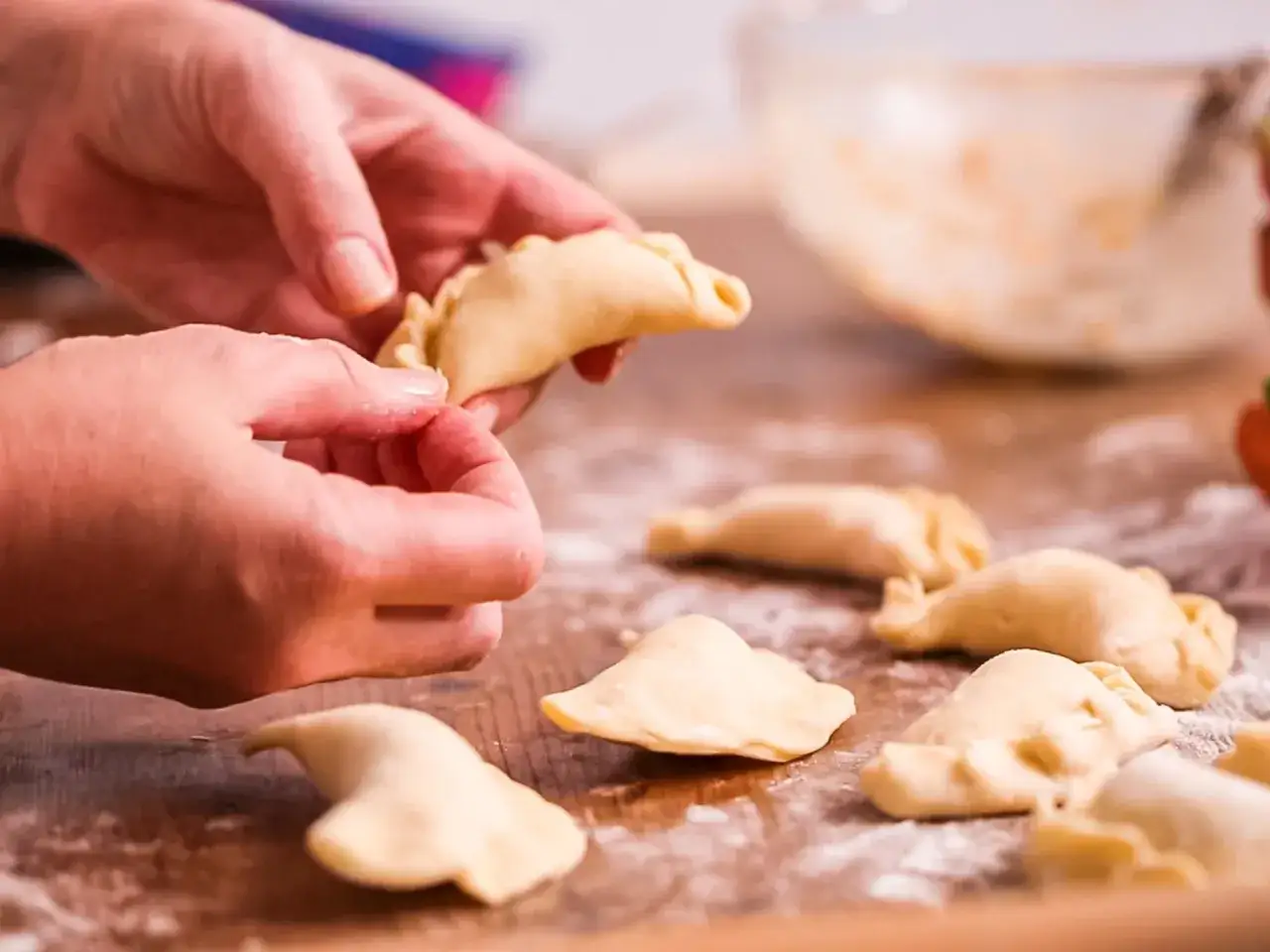Dłonie formują pierogi z serem na drewnianym blacie, obok leżą gotowe, posypane mąką.