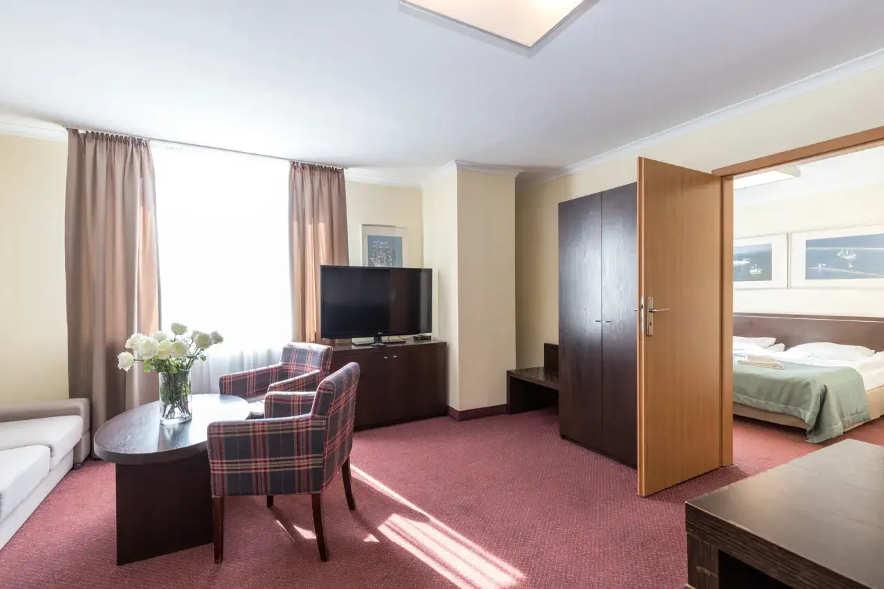Pokój w resort spa plaza: wygodne fotele, stolik z kwiatami, telewizor i drzwi do sypialni.