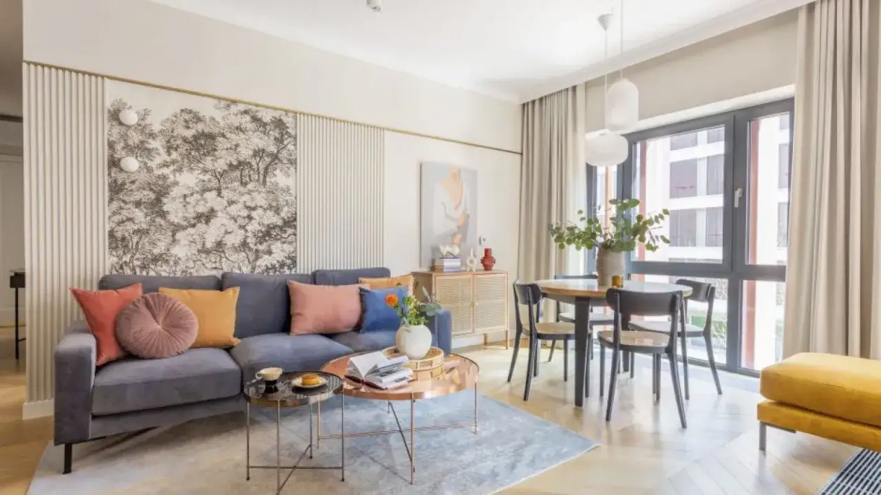 Nowoczesny salon z szarą kanapą i kolorowymi poduszkami. Na ścianie tapeta z drzewami, inspiracja jak powiesić obraz na ścianie.