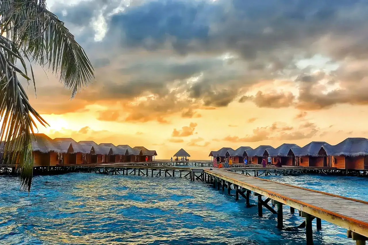 Zachód słońca na Malediwach, idealny czas, by tu przyjechać. Drewniane domki na wodzie, palmy i turkusowa laguna.
