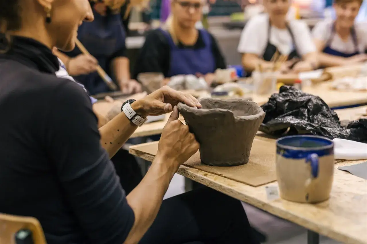 Kreatywne zajęcia z ceramiki. Dłonie formują gliniany dzbanek, w tle inni uczestnicy tworzą swoje dzieła.