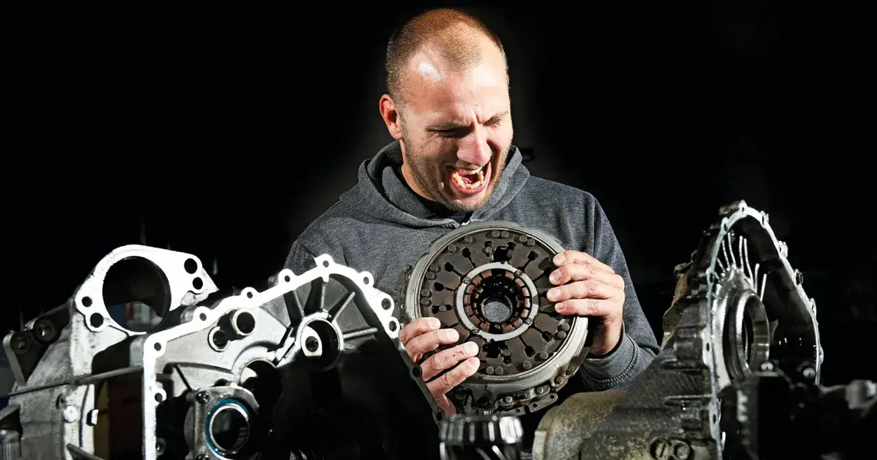 Mężczyzna z grymasem na twarzy trzyma sprzęgło, obok części skrzyni automatycznej DSG.