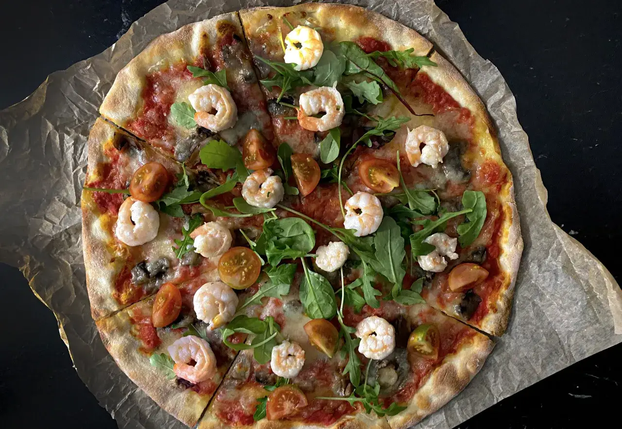 Pyszna pizza z krewetkami, pomidorkami i rukolą, podana na papierze do pieczenia.