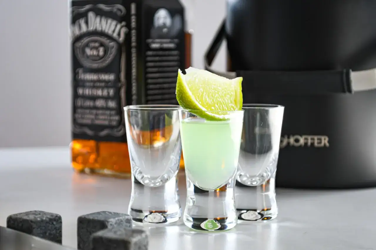 Jak dobrać odpowiedni kieliszek do wódki degustacyjny vs shot? Trzy małe kieliszki, jeden z zielonym drinkiem i limonką, obok butelki Jack Daniel's.