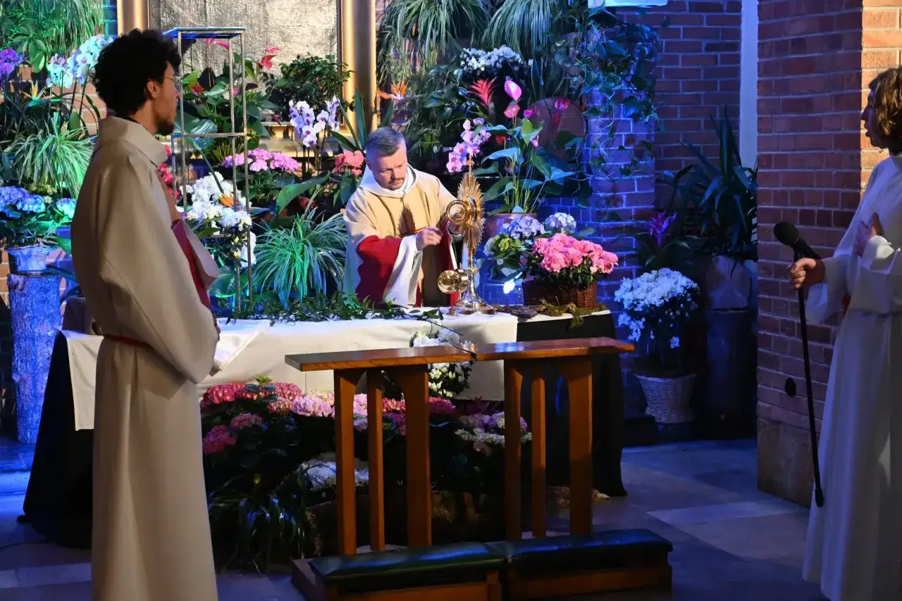 Uroczysta Wigilia Paschalna trwa. Kapłan w szatach liturgicznych adoruje monstrancję wśród bogactwa kwiatów.