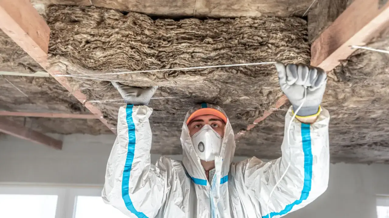 Pracownik w kombinezonie ochronnym montuje wełnę mineralną na suficie. Do precyzyjnego cięcia wełny mineralnej używa specjalnych narzędzi.