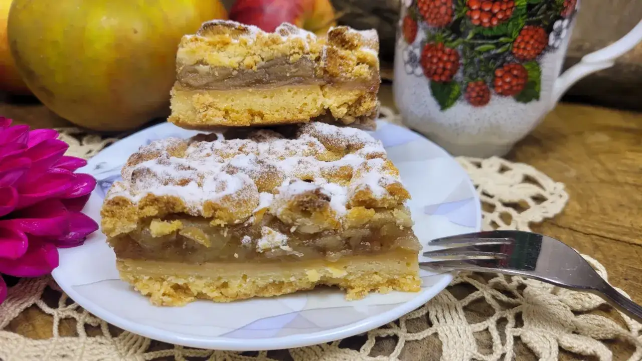 Dwa kawałki szarlotki posypane cukrem pudrem, obok jabłka i kubek z malinami. Zastanawiasz się, ile czasu piec szarlotkę?