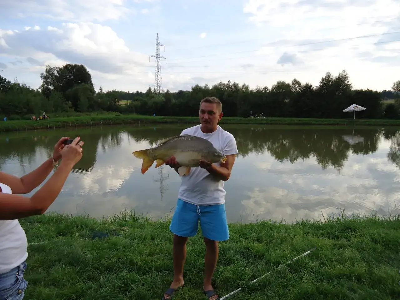 Szukasz miejsca gdzie na ryby bez karty Kraków? Mężczyzna z dumą prezentuje złowionego karpia nad stawem.