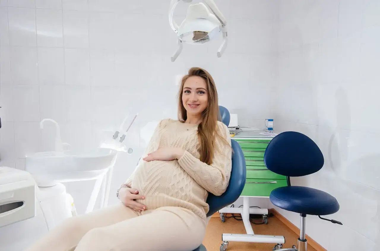 Uśmiechnięta ciężarna kobieta w gabinecie stomatologicznym, przygotowująca się do fluoryzacji. Dba o zdrowie zęb&oacute;w dla siebie i dziecka.