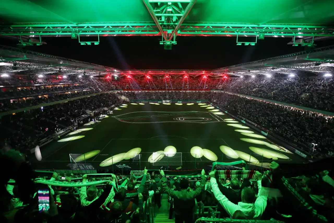 Stadion Legii Warszawa rozświetlony zielonymi i czerwonymi światłami. Tłum kibiców z szalikami, gotowy do dopingu swoich ulubionych klubów piłkarskich w Warszawie.