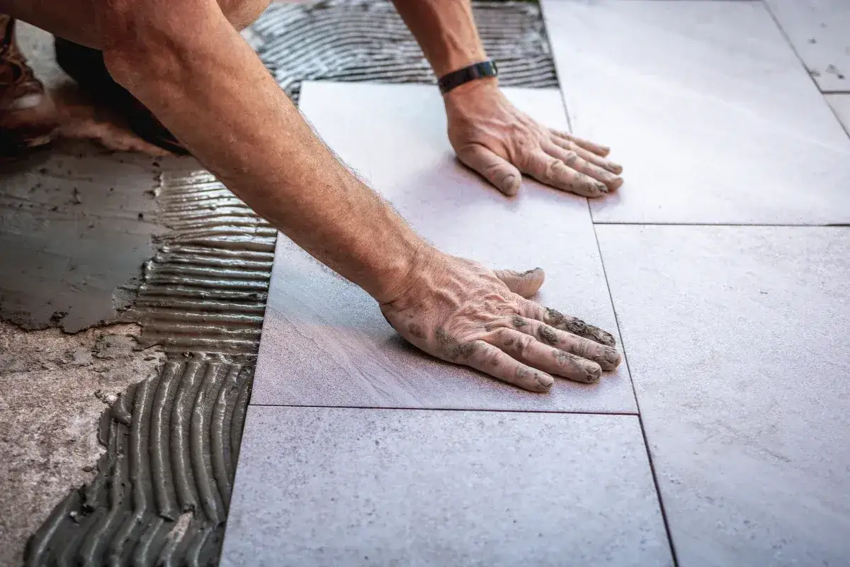 Ręce układają płytki na zaprawie, pokazując dopuszczalną grubość kleju pod płytki.