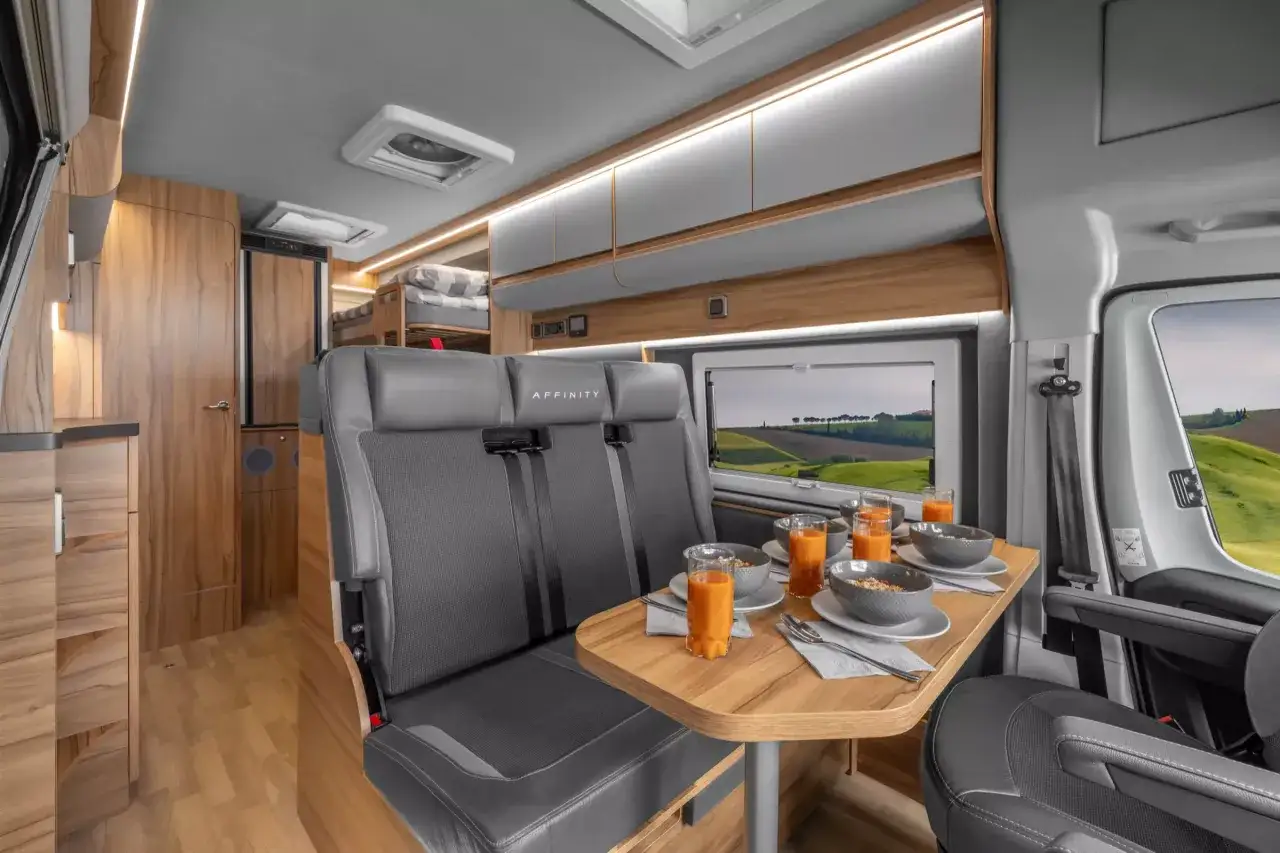 Wnętrze kampera na 5 os&oacute;b z wygodnymi siedzeniami, stołem i widokiem na zielone wzg&oacute;rza.