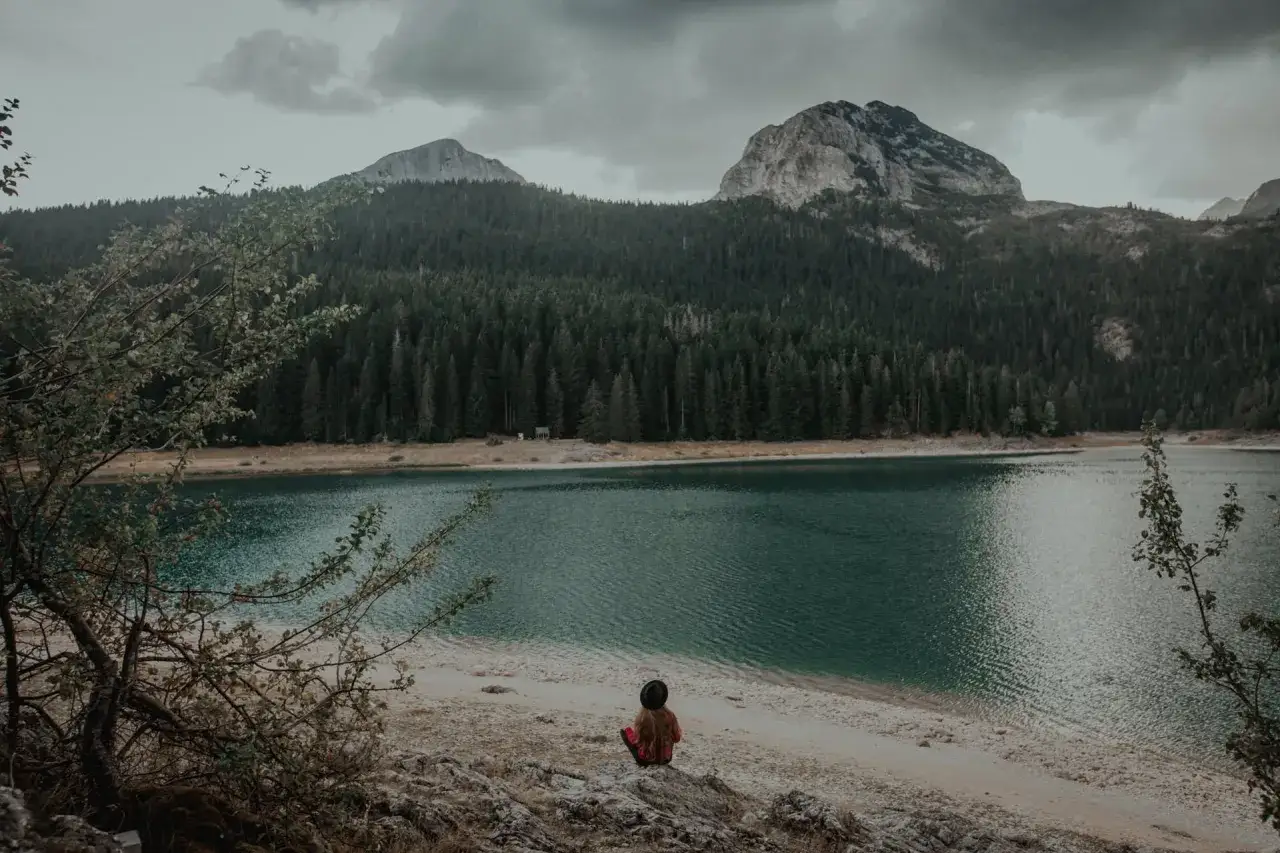 Dziewczynka w czarnym kapeluszu siedzi na brzegu malowniczego jeziora w Czarnog&oacute;rze, otoczona lasem i g&oacute;rami.