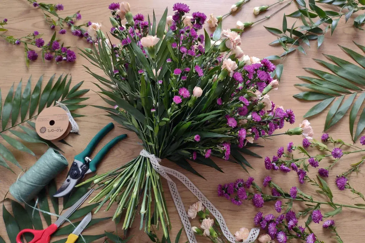 R&oacute;żnorodne rodzaje bukiet&oacute;w we florystyce: bukiet z fioletowych i brzoskwiniowych kwiat&oacute;w, narzędzia florystyczne i zielone liście.
