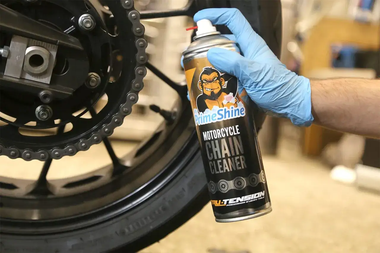 Rękawiczka w niebieskiej rękawicy trzyma puszkę z preparatem do smarowania łańcucha motocyklowego.