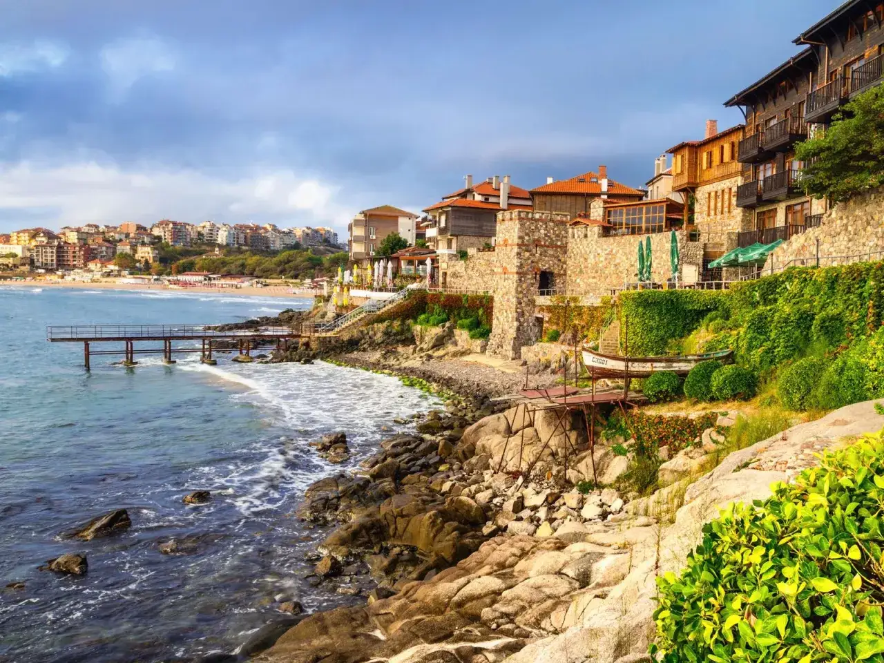 Kamieniste wybrzeże z budynkami i plażą w tle. Zastanawiasz się, ile kosztuje lot do Bułgarii?