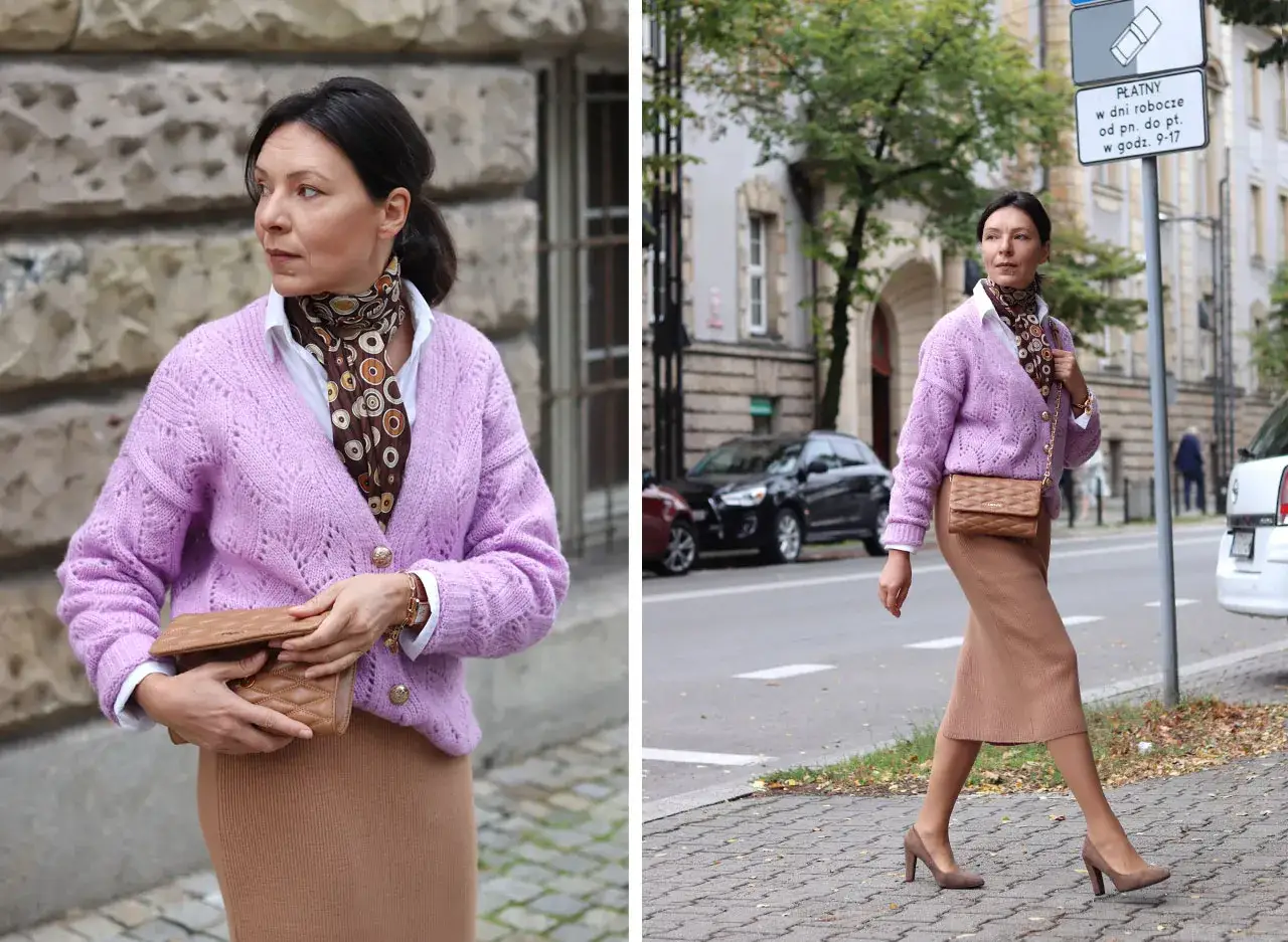 Elegancka kobieta w swetrze w kolorze liliowym i brązowej sp&oacute;dnicy, trzymająca torebkę.