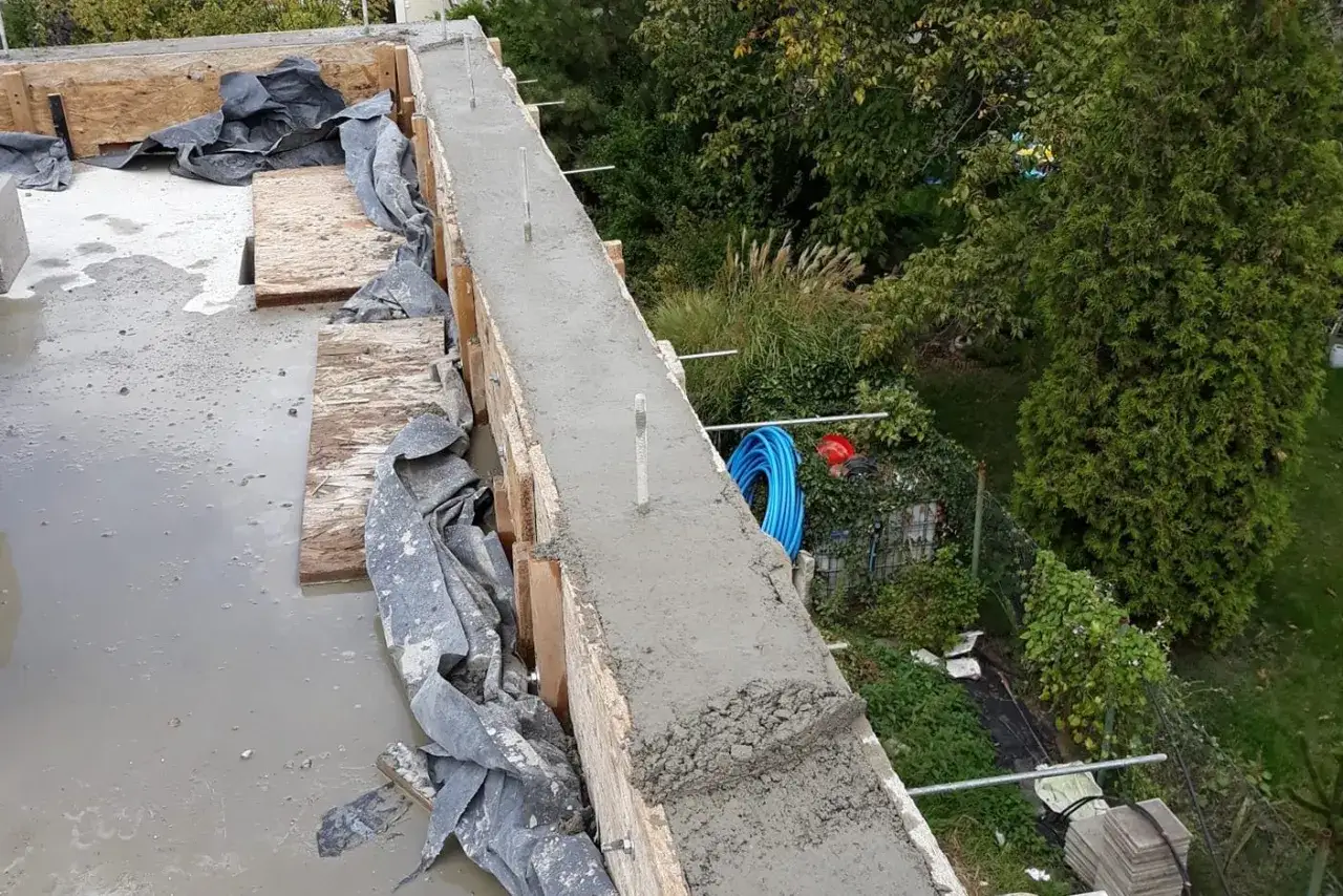 Świeży beton na stropie, widać szalunki i kilka szpilek do murlaty. W tle zieleń i narzędzia budowlane.