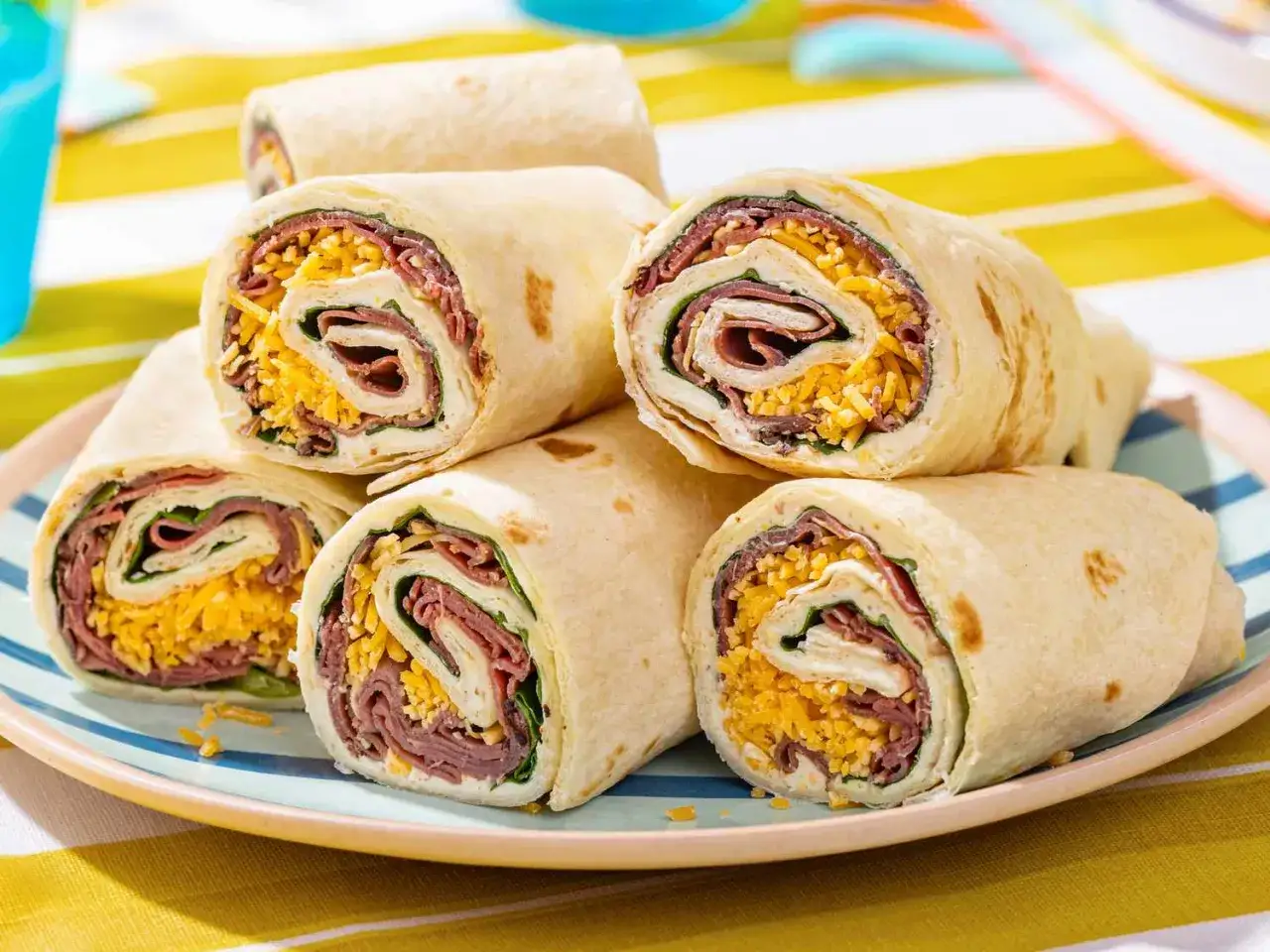 Sześć pysznych wrapów z tortilli, wypełnionych serem, wędliną i zielonymi liśćmi, ułożonych na talerzu.