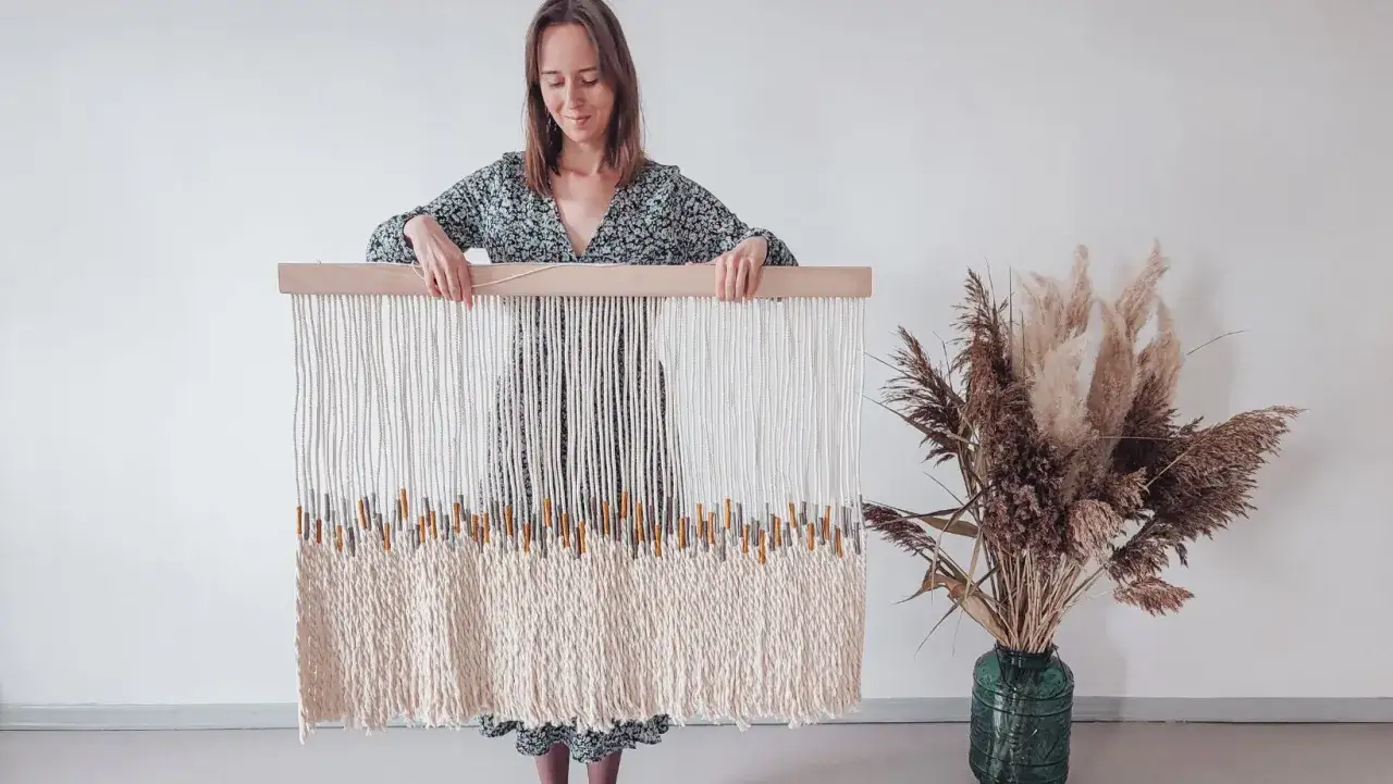 Kobieta prezentuje swoje dzieło – sztukę plecenia sznurków. Makrama z drewnianymi elementami i ozdobnymi pęczkami traw.