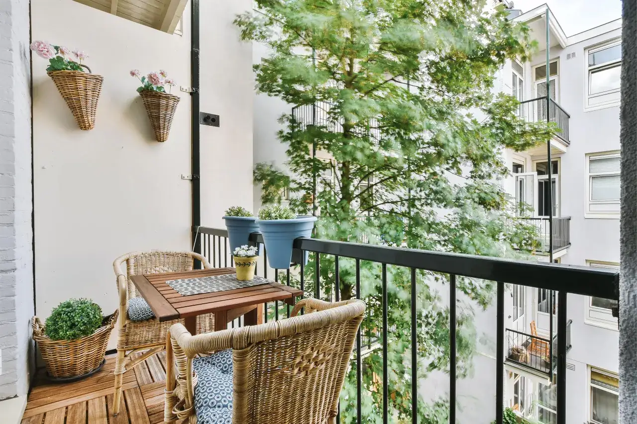 Przytulny mini balkon w bloku z wiklinowymi meblami, kwiatami w koszach i widokiem na zielone drzewa.
