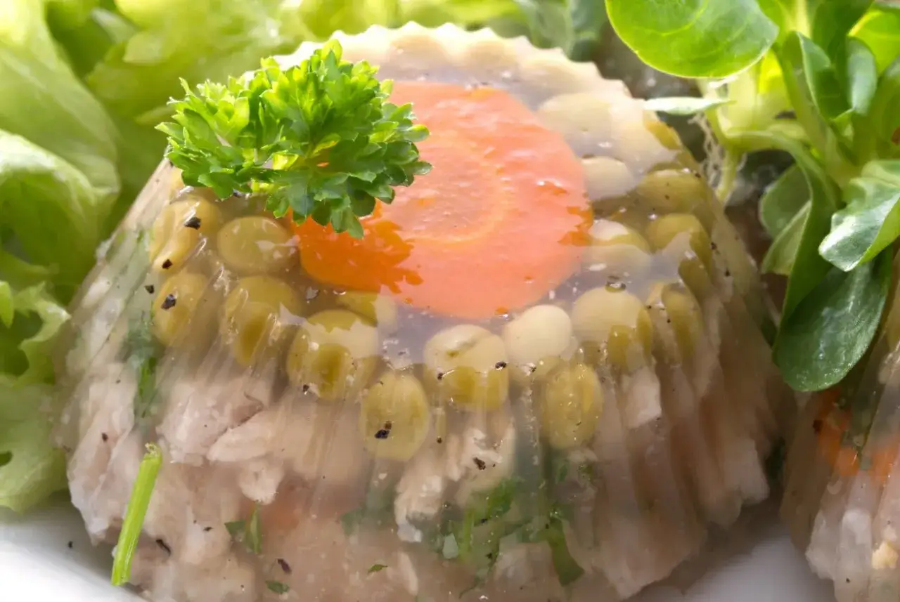 Jak zrobić żelatynę do garmażerki? Zobacz, jak apetycznie wygląda galareta z mięsem, groszkiem i marchewką, ozdobiona natką pietruszki.