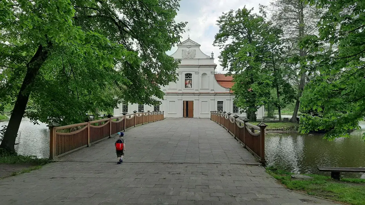 Most na wodzie prowadzi do białego kościoła na Zwierzyńcu. Dziecko z plecakiem idzie w stronę świątyni.