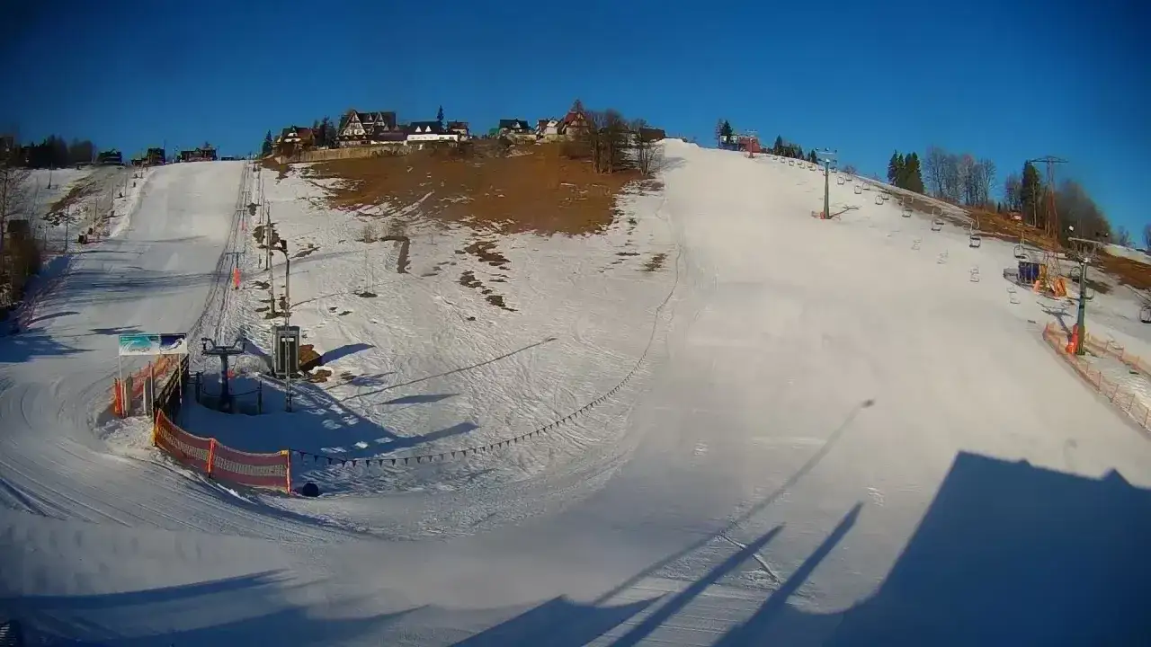 Słoneczny dzień na olczańskim wierchu. Stok narciarski z wyciągiem krzesełkowym i kilkoma domkami w tle.