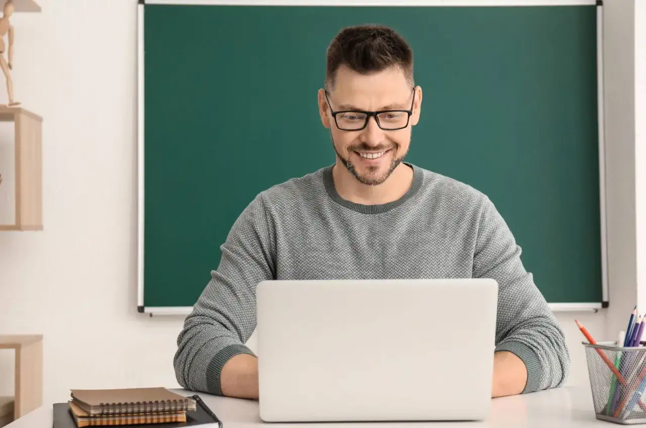 Uśmiechnięty nauczyciel w okularach pracuje przy laptopie. Czekamy na laptopy dla nauczycieli, by usprawnić pracę.