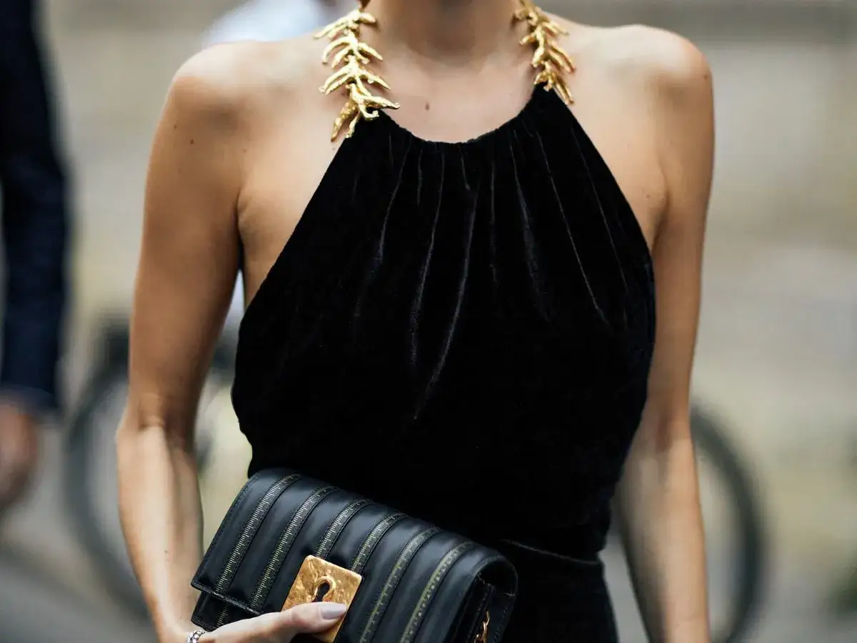 Elegancka czarna sukienka z aksamitu z dekoltem typu halter i złotą biżuterią na szyi. Kobieta trzyma czarną kopert&oacute;wkę.