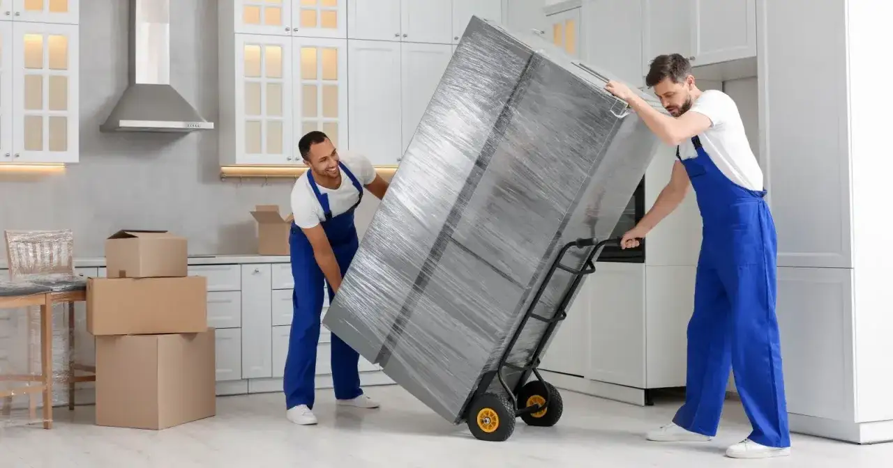 Dw&oacute;ch mężczyzn w niebieskich kombinezonach przewozi lod&oacute;wkę na w&oacute;zku. To dobry przykład, jak przewieźć lod&oacute;wkę bezpiecznie.