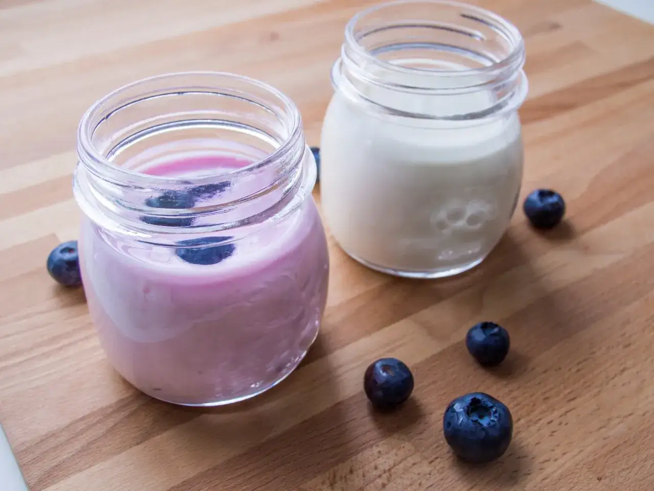 Dwa słoiczki z jogurtem, jeden r&oacute;żowy z jagodami, drugi biały. Wok&oacute;ł świeże bor&oacute;wki. Wegańskie kultury bakterii jogurtowych co to jest?