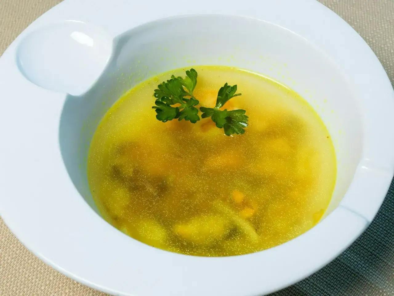 Złocista zupa na chorobę, podana w białej miseczce, ozdobiona gałązką natki pietruszki. Idealna na przeziębienie.