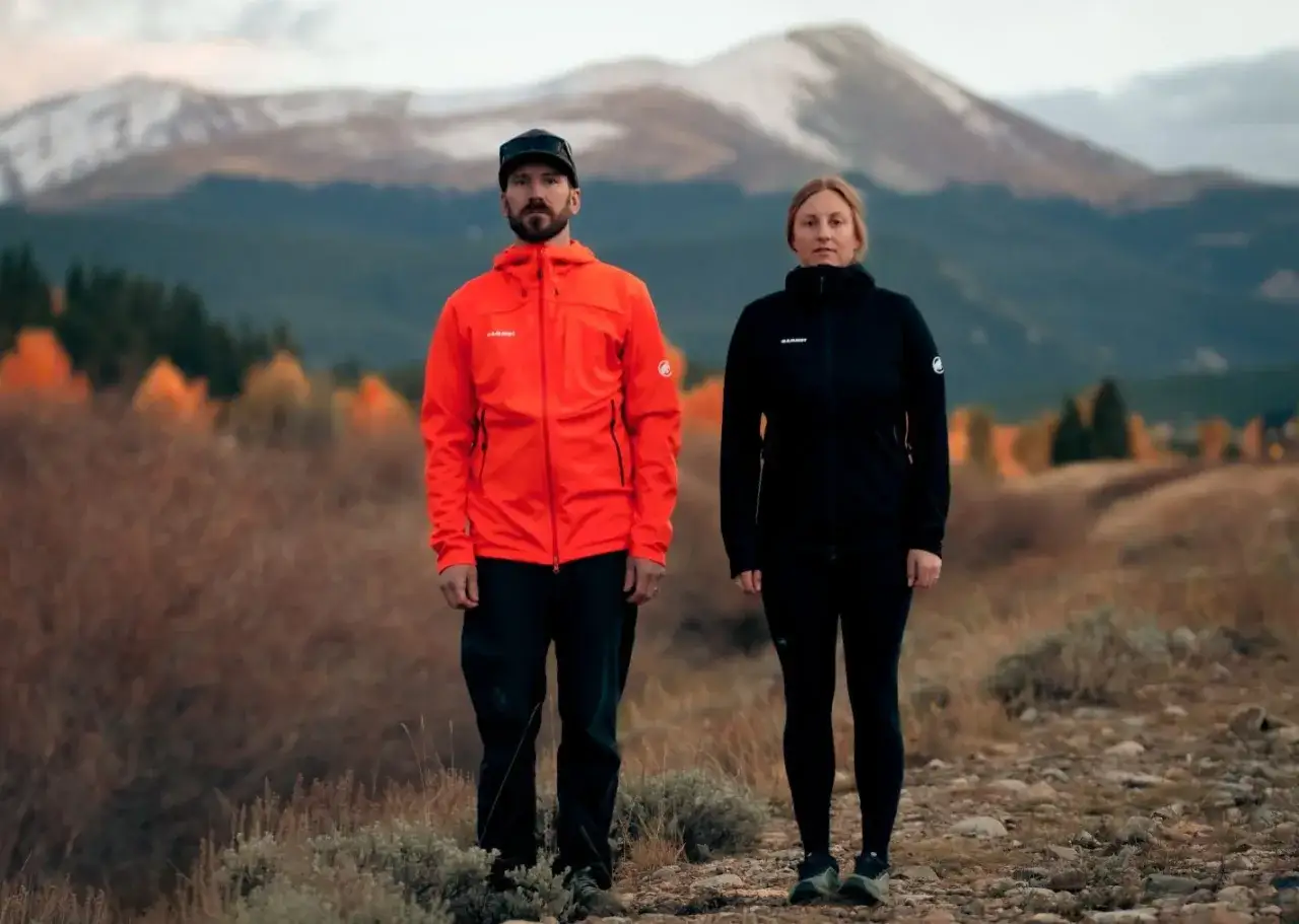 Para, ubrana w pomarańczowy softshell, i kobieta w czarnym, stoją na tle gór. To idealny przykład, co to softshell - lekka, oddychająca odzież na wędrówki.
