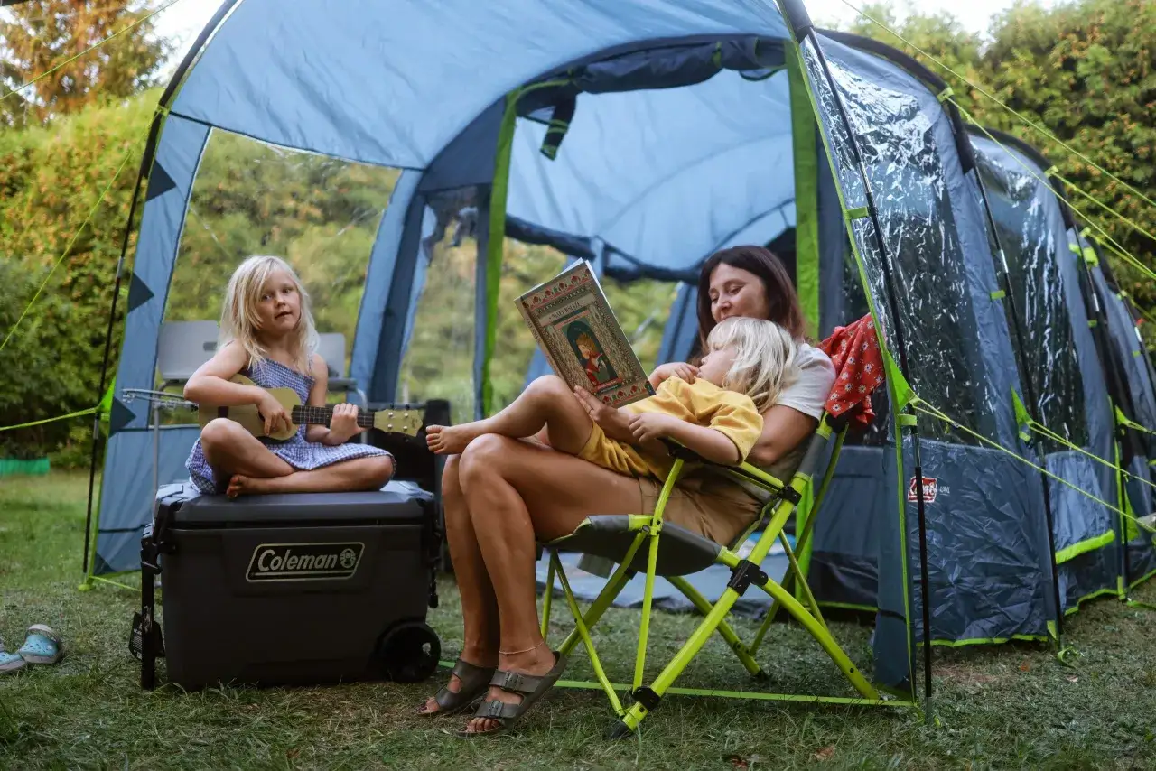 Mama czyta dzieciom bajkę pod namiotem, a starsza córka gra na ukulele. To idealny przykład, co zabrać pod namiot z dziećmi: książki i instrumenty.
