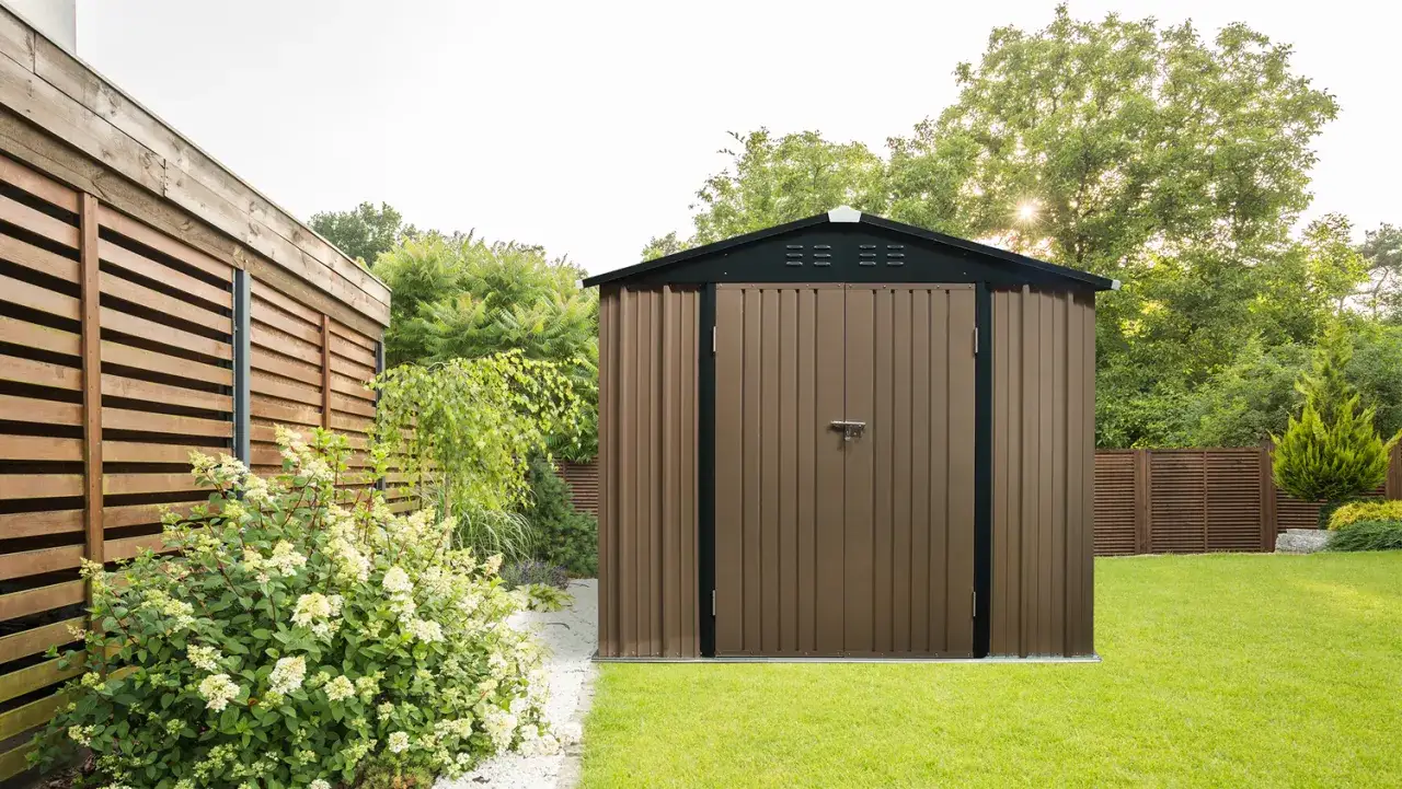 Brązowy domek na narzędzia ogrodowe stoi na zielonym trawniku, obok drewnianego ogrodzenia i kwitnących krzewów.