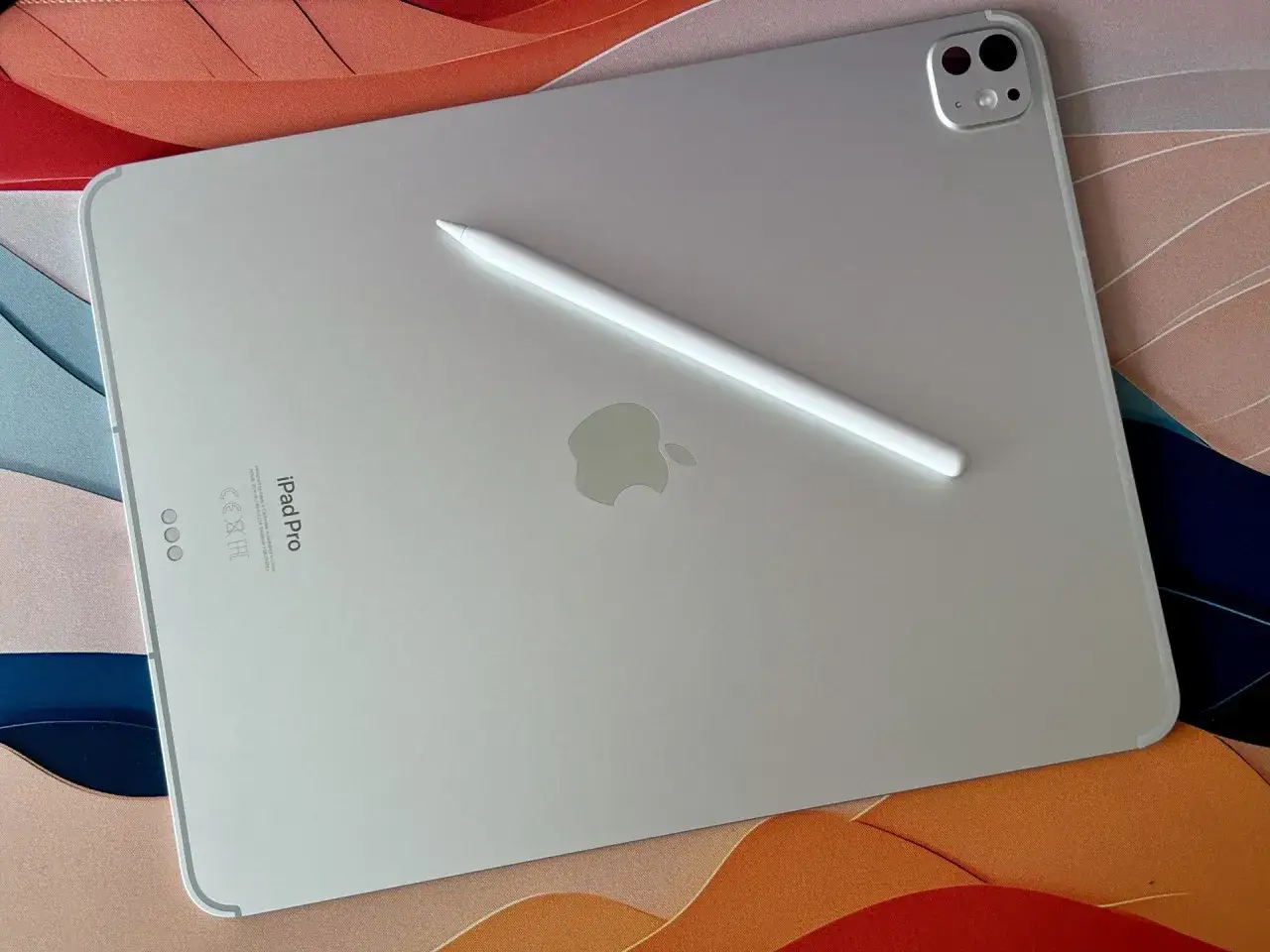 Srebrny iPad Pro z Apple Pencil na tle kolorowej podkładki. Zastanawiasz się, cellular co to? To wersja z łącznością kom&oacute;rkową.