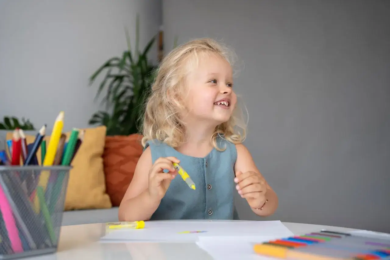 Uśmiechnięta dziewczynka z blond włosami bawi się kredkami, ćwicząc gry i zabawy językowe w języku polskim.