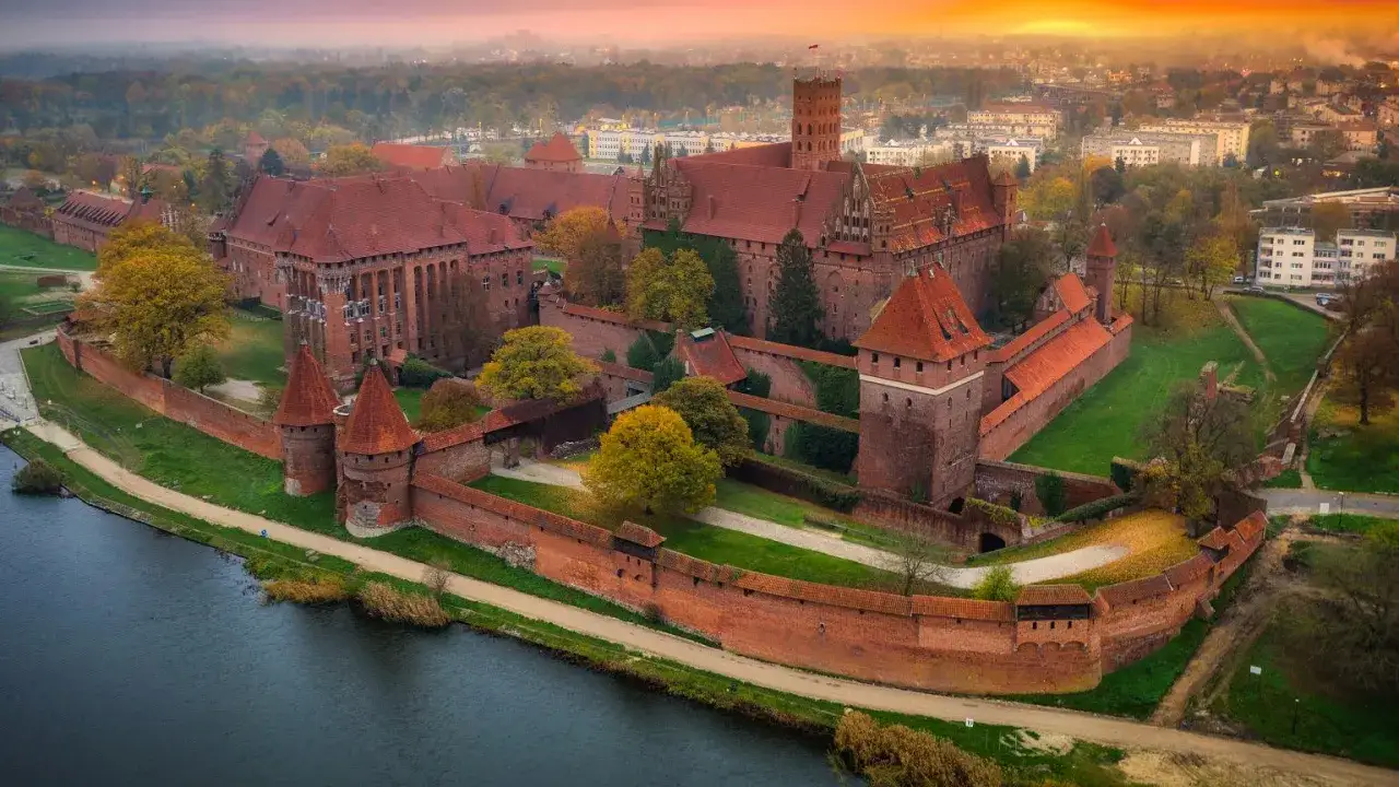 Zamek w Malborku, gdzie nagrywano Krzyżaków, w jesiennej scenerii o zachodzie słońca. Potężne ceglane mury i wieże otaczają rozległy kompleks.