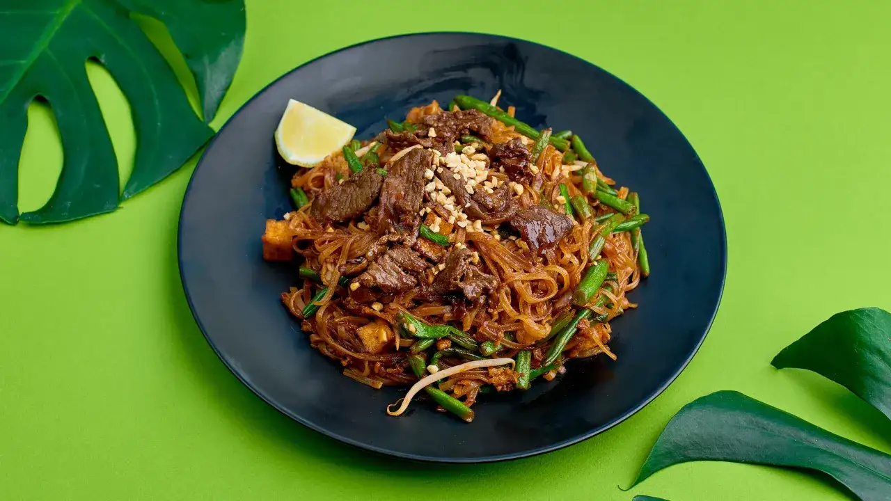 Pyszny Pad Thai przepis z wołowiną, fasolką szparagową i orzeszkami ziemnymi na czarnym talerzu, ozdobiony liśćmi monstery.
