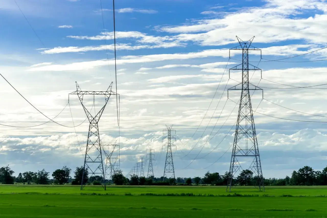 Linia energetyczna w trakcie likwidacji. Wysokie słupy energetyczne na tle błękitnego nieba z chmurami, nad zielonym polem.