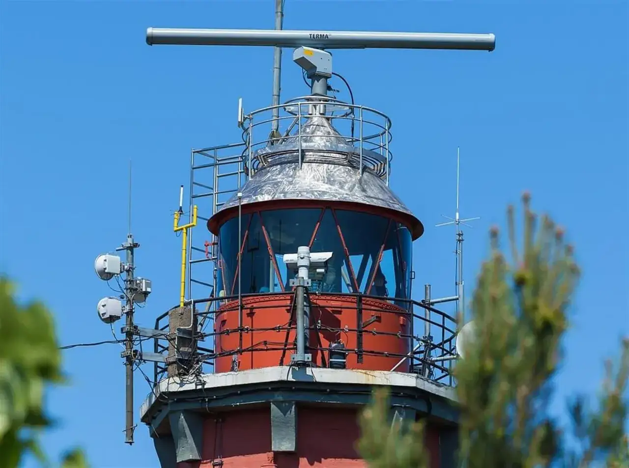 Czerwona latarnia morska Hel z metalową kopułą i radarem na szczycie, otoczona antenami i zielenią.