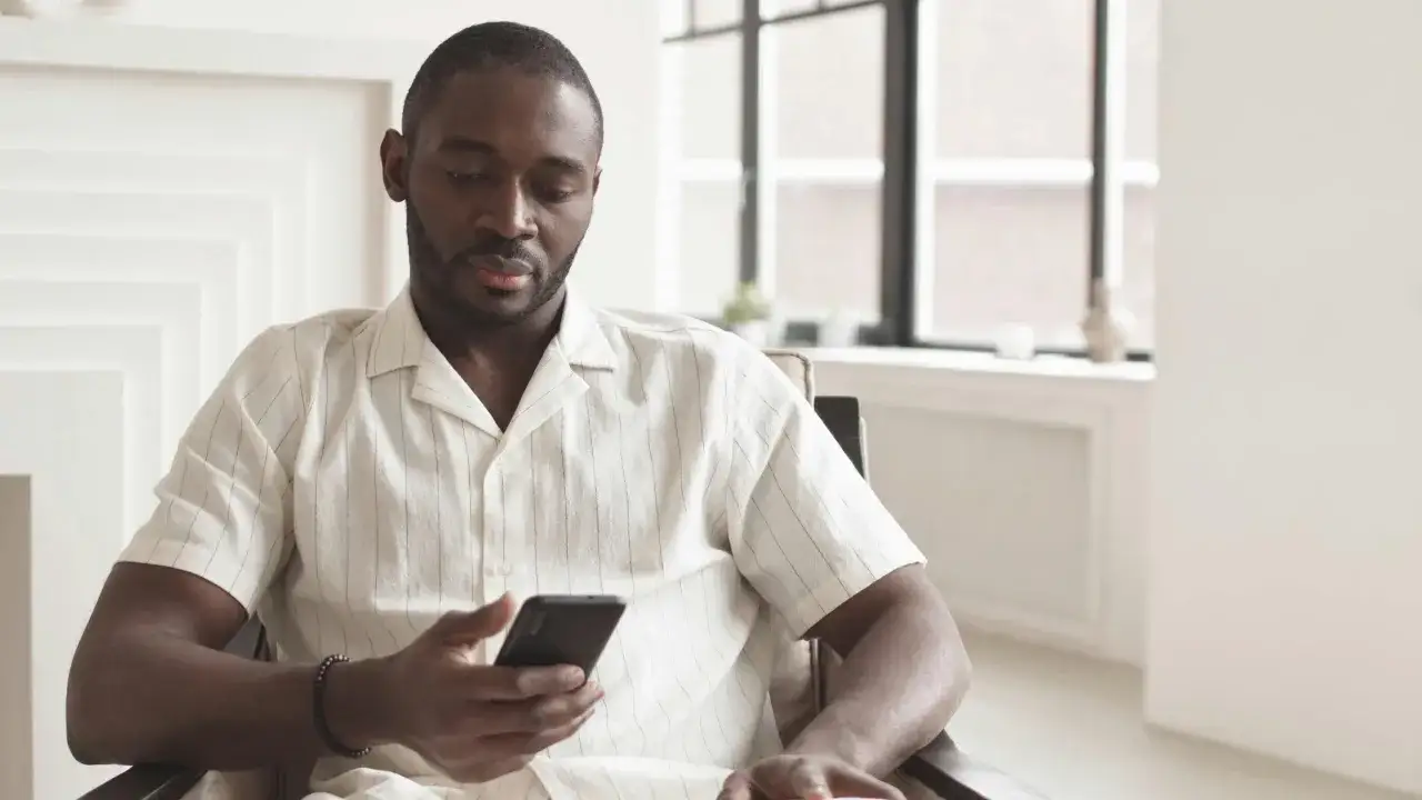 Mężczyzna w jasnej koszuli siedzi i patrzy na telefon, być może właśnie sprawdza, co to jest meet w telefonie, planując spotkanie.
