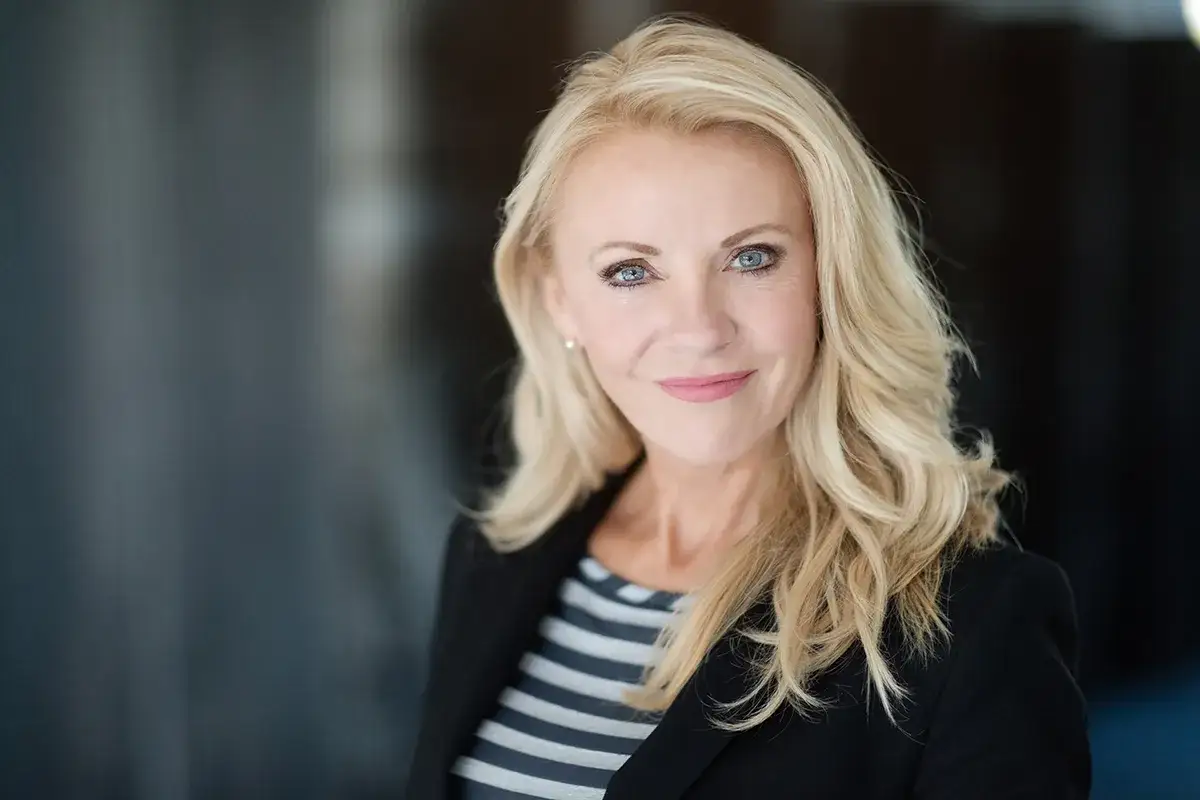 Uśmiechnięta blondynka w marynarce, z subtelnym makijażem biznesowym do zdjęć, prezentuje profesjonalny wizerunek.